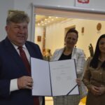 Na obrazku trzy osoby w pomieszczeniu, które się pojawiają. Jeden mężczyzna trzyma otwarty dokument z czerwoną pieczęcią, a po jego bokach dwie kobiety - cała scena ma profesjonalny charakter.