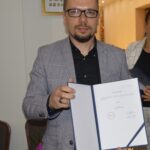 Na zdjęciu jest mężczyzna w okularach i szarym garniturze, który trzyma otwartą teczkę z dokumentami, przechowywaną w pomieszczeniu. Wydaje się być skupionym nad tym, co jest na kartach.