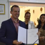 Na widok mężczyzn w garniturze, trzymającego dokumentu, który stoi i się uśmiecha w biurze z dwiema kobietami. Jedna z kobiet bije brawo.