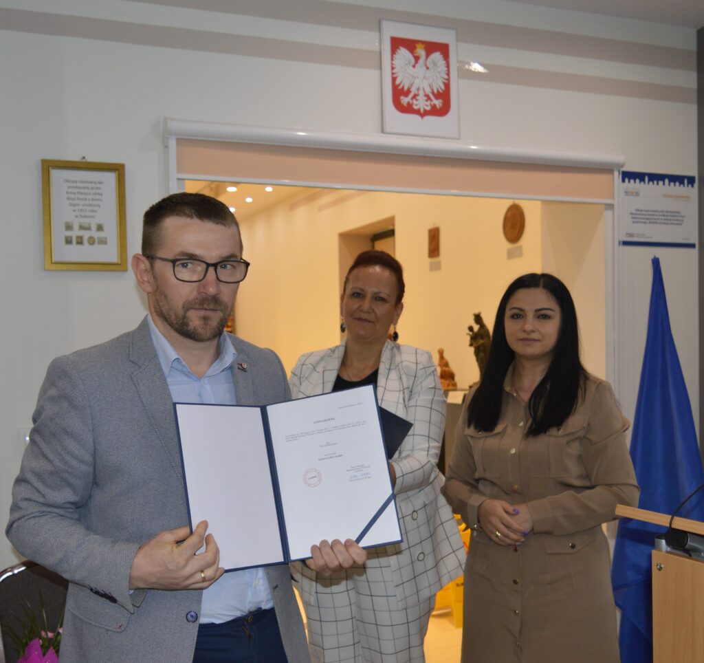 Na obrazku znajdują się trzy osoby - dwie kobiety i jeden mężczyzna, którzy są objęci certyfikatem. W tle widoczne są godło Polski i flagi Unii Europejskiej.