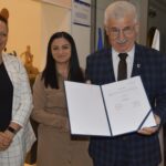 Na obrazku dwie kobiety i mężczyźni w środowisku biznesowym. Trzyma otwarty dokument, a za nim znajduje się płyta z ceramiką.