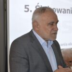 Na obrazku dojrzałego mężczyzn o siwych włosach i brodzie, przemawiającego z mównicy. Jest ubrany w szarą koszulę i stoi przed prezentacją.