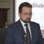 Na obrazku widoczny jest mężczyzna w niebieskim garniturze i okularach, który przemawia do mikrofonu przy mównicy. W tle zauważalny jest ekran prezentacyjny.