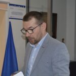 Na widok mężczyzn w okularach i szarych marynarkach, który przemawia przy podium z mikrofonami. W tle znajdują się flagi Unii Europejskiej oraz różne artefakty.