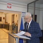 Na obrazku starszego mężczyznę o szarych włosach, noszącego okulary i garnitur, który czyta z dowodu przy podium z mikrofonem i flagami narodowymi. Cała scena rozgrywająca się w muzeum.