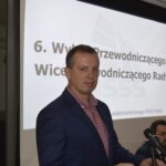 Na obrazku mężczyzn w niebieskim garniturze i czerwonym krawacie, który stoi przed mikrofonem i przemawia do głośnika. W tle jest prezentacja slajdu.