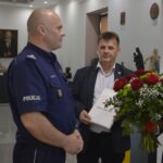 Na obrazku dwóch mężczyzn stojących w pomieszczeniu; jeden z nich nosi mundur policyjny i trzyma ręce razem, a drugi w garniturze trzyma zielone czerwono-białych kwiatów i zwierząt z prezentem. W tle widać wystawione dzieło sztuki.