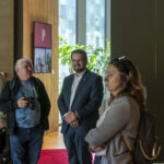 DSC_4232 Na obrazku znajdują się trzy osoby, które są dobrze oświetlone, rozmawiają ze sobą. Jedna z nich trzyma urządzenie fotograficzne, a w tle jest duże okno oraz roślina.