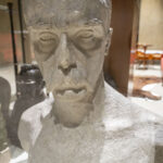 DSC_4250 Na obrazku pojawia się zbliżenie na kamienne popiersie przedstawiające osobę o długich włosach, wąsach i wyrazistych rysach twarzy, umieszczonych w galerii. Tło oznaczone na muzealnej oprawie eksponatu, przeznaczonej do stosowania w charakterze artystycznym dzieła.