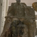 DSC_4266 Na miejscu pojawia się brązowa rzeźba siedząca jako postać z zamyśloną miną, ustawiona na półce wystawowej, której widoczne są inne artefakty w tle. Otoczenie ograniczone przestrzenią muzealną lub galerią sztuki.