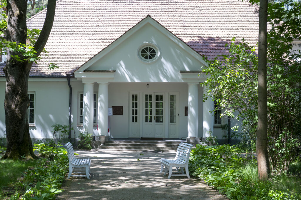 Na miejscu pojawia się biały dom z dwuspadowym dachem, dostępny z drzewami i zielenią. Na ścieżce do wejścia są dwie białe ławki.