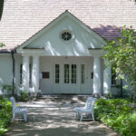 DSC_4306 Na miejscu pojawia się biały dom z dwuspadowym dachem, dostępny z drzewami i zielenią. Na ścieżce do wejścia są dwie białe ławki.