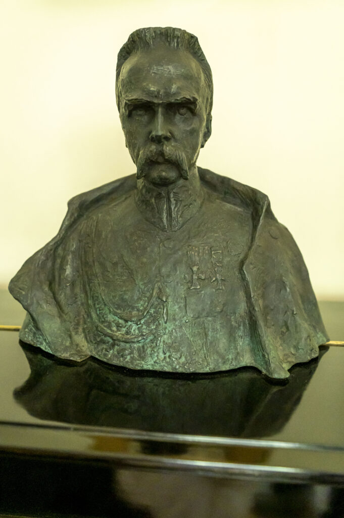 Na obrazku pojawia się popiersie mężczyzny z wąsami, ubranego w szczegółowo wyrzeźbiony mundur, który jest wyświetlany jako medale. Popiersie jest eksponowane na tle jednolitego tła.