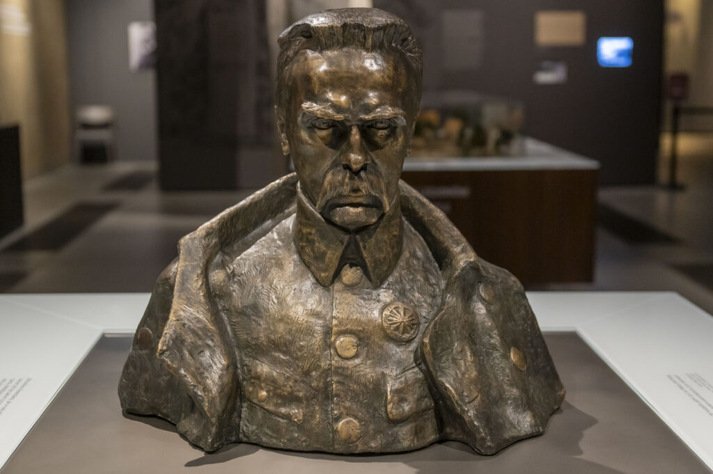 Na obrazku pojawia się popiersie mężczyzny o wyrazie twarzy, ubranego w mundurze wojskowym. Rzeźba jest eksponowana w muzeum, co następuje neutralne tło i profesjonalne oświetlenie.