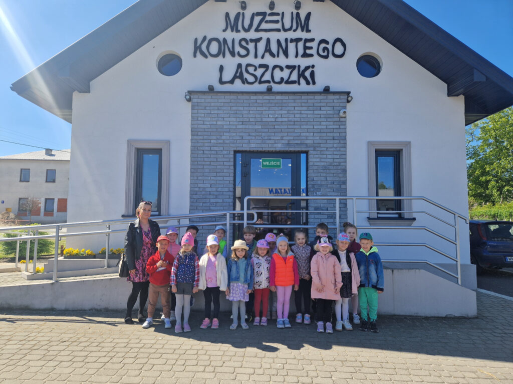 Na wolności jest grupa dzieci oraz dorosła osoba przed wejściem do „Muzeum Konstantego Laszczyka”. Tło plastikowe przezroczyste, błękitne niebo.