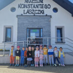 Na obrazku grupa dzieci stoi przed wejściem do Muzeum Konstantego Laszczki, wejście z pogodnego słońca dnia. Dzieci wyglądają na podekscytowane i gotowe na zwiedzanie, otoczone zielenią i ciepłym światłem słońca.