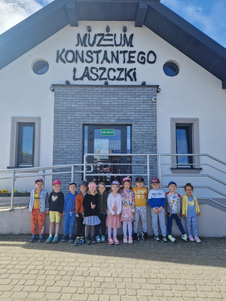 Na obrazku grupa dzieci stoi przed wejściem do Muzeum Konstantego Laszczki, wejście z pogodnego słońca dnia. Dzieci wyglądają na podekscytowane i gotowe na zwiedzanie, otoczone zielenią i ciepłym światłem słońca.