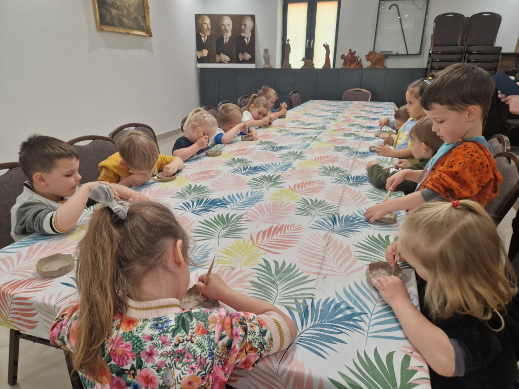 Dzieci siedzą przy stole, zaangażowane w obowiązki zawodowe z gliny pod dorosłymi. Stół pokryty jest kolorowym obrusem w liściastym wzorze, a w tle są portrety.