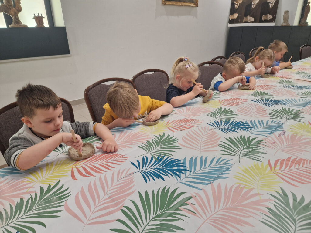 Na obrazku dzieci siedzące przy długim stole, który jest przykryty kolorowym obrusem w liściach, każde z nich trzyma pędzel i pracuje nad rozwiązaniem projektami z tworzyw sztucznych. Atmosfera jest pełna skupienia i aktywności, co ochronne, wspólne zajęcia artystyczne.