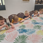 Na obrazku dzieci siedzące przy długim stole, który jest przykryty kolorowym obrusem w liściach, każde z nich trzyma pędzel i pracuje nad rozwiązaniem projektami z tworzyw sztucznych. Atmosfera jest pełna skupienia i aktywności, co ochronne, wspólne zajęcia artystyczne.