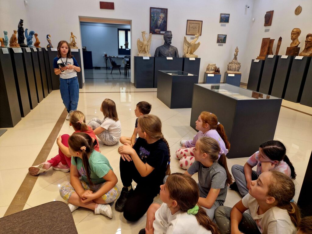 Na ekranie pojawia się grupa dzieci siedzących na słuchawkach, które słuchają dziewcząt, które stoją i słuchają. Scena ma miejsce w galerii sztuki pełnych rzeźb i popiersi zawartych na postumentach.