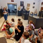 Na ekranie pojawia się grupa dzieci siedzących na słuchawkach, które słuchają dziewcząt, które stoją i słuchają. Scena ma miejsce w galerii sztuki pełnych rzeźb i popiersi zawartych na postumentach.