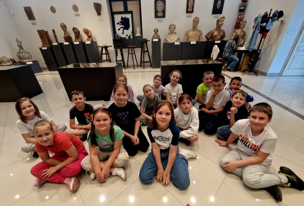 Grupa dzieci siedzi na miejscu w muzeum, z rzeźbami i popiersiami na postumentach za nimi. Otoczenie szczególne edukacyjne lub kulturalne dla wstępnego w organizmie artystycznym.