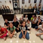 Grupa dzieci siedzi na miejscu w muzeum, z rzeźbami i popiersiami na postumentach za nimi. Otoczenie szczególne edukacyjne lub kulturalne dla wstępnego w organizmie artystycznym.