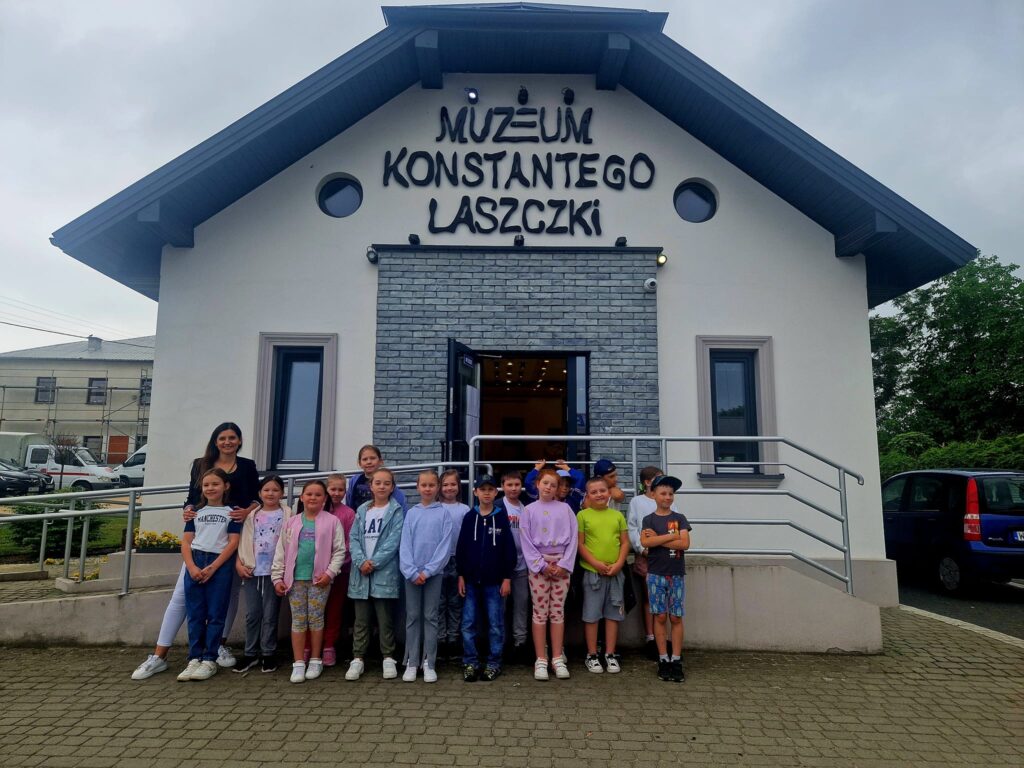 Na obrazku grupy dzieci oraz jednego dorosłego, którzy pozują do zdjęć przed wbudowanym z szyldem "Muzeum Konstantiego Laszczki". W tle jest fasada muzeum poświęconego twórczości rzeźbiarza Konstantina Laszczki.