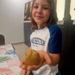 Na obrazku dziecka z długimi włosami, które znajdują się w pomieszczeniu i trzymają małe gliniane rzeźby. Dziecko ubrane jest w biało-niebieskiej wersji z napisem „Manchester” i uśmiecha się szeroko.