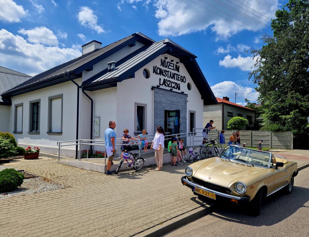 Na obecność grupy ludzi, w tym dzieci z rowerami, należy przed Muzeum Konstantego Laszczki. Na pierwszym planie jest beżowy kabriolet, całość rozgrywająca się w słonecznym dniu z błękitnym niebem i chmurami.