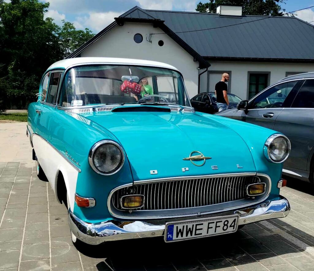 Na obrazku widnieje zabytkowy niebiesko-biały samochód marki Opel wprowadzonyny na podjeździe. W tle można dotrzeć do domu oraz innych samochodów.