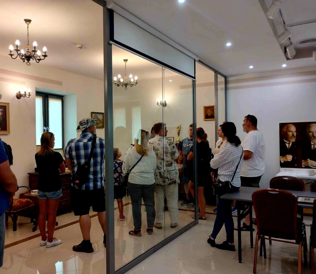 Grupa ludzi w zwykłych strojach stoi w muzealnej sali, oglądając eksponat za szklaną przegrodą. Na ścianach widzisz różne obrazy, a pokój znajduje się w zabytkowych meblach.