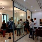 Grupa ludzi w zwykłych strojach stoi w muzealnej sali, oglądając eksponat za szklaną przegrodą. Na ścianach widzisz różne obrazy, a pokój znajduje się w zabytkowych meblach.