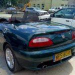 Na obrazku pojawia się ciemnozielony kabriolet z czarnym miękkim dachem i żółtą tablicą rejestracyjną, wprowadzonyny na wybrukowanym placu. W tle widoczne są inne bagaże oraz budynek.