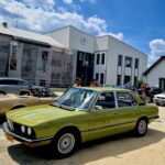 Ludzie zgromadzeni są wokół zabytkowych samochodów, w tym zielonym sedana, specyficznego kabrioletu oraz srebrnego hatchbacka, na słonecznym terenie z dodatkowymi i zbudowanymi budynkami w tle. Otoczenie na wydarzenie wydarzenia plenerowego poświęconego motoryzacji.