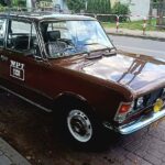 Na ulicy znajduje się zabytkowy brązowy samochód z naklejką „WPT 1313”, umieszczony na ulicy, wyposażony w żółte światła przeciwmgielne oraz znak TAXI na dachu. W tle widać srebrny samochód oraz chodnik drzewami.