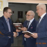 Na obrazku trzech mężczyzn w garniturach wymieniających uściski dłoni oraz trzyma plakiety, co ważne formalną lub wręczenie nagród. W odpowiednim dolnym rogu widnieje dane „27.03.2024” i godzina „14:24”, wydarzenie moment tego wydarzenia.