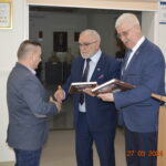 Na obrazku trzech mężczyzn ubranych w ubrania biznesowe wewnątrz pomieszczenia; jeden z nich nosi się i ściska drugiemu, trzymając przy tym oprawiony dokument. Trzeci mężczyzna trzyma oprawiony dokument, a w tle są podium oraz plakietka na ścianie.