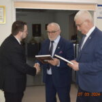 Na obrazku znajdującym się trzech mężczyzn w garniturach w formalnym otoczeniu. Dwóch z nich trzyma twardookładkowe książki, a jeden z nich ściska dłoń trzeciegomu mężczyźnie, przy czym w rogu jest data i godzina „27.03.2024 14:25”.