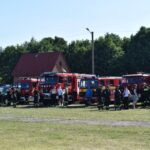 Na ekranie pojawia się kilka wozów strażackich podłączonych w szeregu z charakterystycznymi i cywilnymi stojącymi przed nimi, które mogą wystąpić na trawiastym terenie na zewnątrz w pogodnym dniu. W tle widoczne są drzewa oraz budynek.