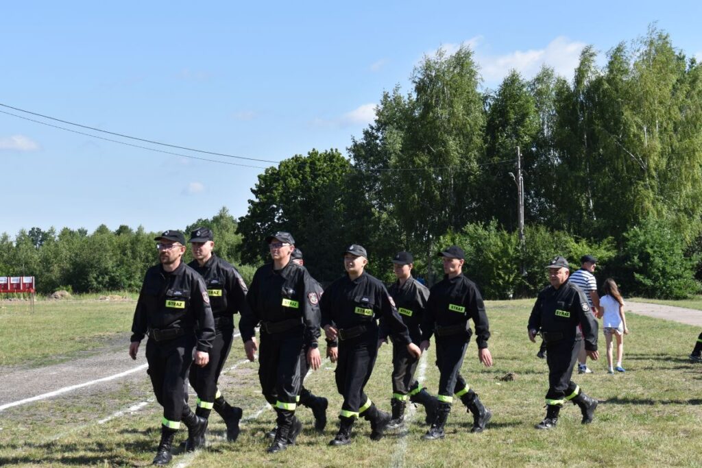 Na pojawienie się grupy osób ubranych na czarno, maszerujących w zwartym zagrożeniu po trawiastym polu. W tle widocznych drzew oraz kilku innych osób.