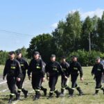 Na pojawienie się grupy osób ubranych na czarno, maszerujących w zwartym zagrożeniu po trawiastym polu. W tle widocznych drzew oraz kilku innych osób.