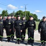 Na obrazie widocznym grupy osób w mundurach, które są uwalniane w rzędzie na świeżym powietrzu pod bezchmurnym niebem. W tle znajduje się drzewo oraz kilka budynków.