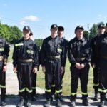 Na zewnątrz znajduje się grupa sześciu mężczyzn w mundurach, uwolniona na zewnątrz w słoneczny dzień. Czterech z nich ma na głowach czapki, cała grupa skonfigurowana jest w szeregu na betonowej powierzchni, która jest widoczna są trawa i drzewo.