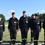 Na sześć osób ubranych w czarne mundury z odblaskowymi pasami stoi w rzędzie na świeżym powietrzu w słoneczny dzień. Wygląda na to, że jest to sygnał bezpieczeństwa lub ratownictwa, gotowego do działania.