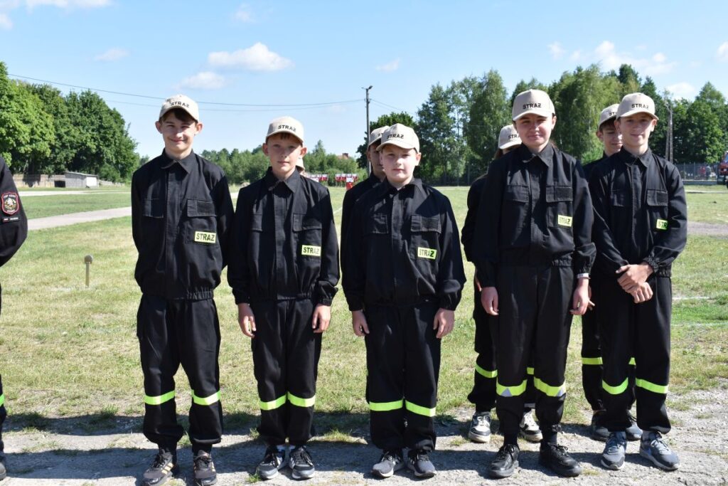 Na obrazie widać grupę młodych osób w czarnych mundurach i beżowych czapkach, które są uwalniane w szeregach na świeżym powietrzu w słoneczny dzień. W tle widać drzewa i roślinność.