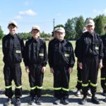 Na obrazie widać grupę młodych osób w czarnych mundurach i beżowych czapkach, które są uwalniane w szeregach na świeżym powietrzu w słoneczny dzień. W tle widać drzewa i roślinność.