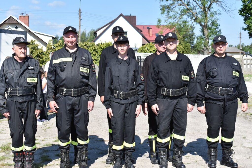 Na obrazku pojawia się grupa sześciu osób ubranych w czarne mundury strażackie z naszymiywkami „STRAŻ”, stojącymi w szeregu na świeżym powietrzu. Część z nich ma na głowach czapki, a za nimi widoczne są okna mieszkalne.