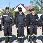 Na obrazku pojawia się grupa sześciu osób ubranych w czarne mundury strażackie z naszymiywkami „STRAŻ”, stojącymi w szeregu na świeżym powietrzu. Część z nich ma na głowach czapki, a za nimi widoczne są okna mieszkalne.